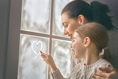 girl drawing a heart on frozen window with her mum in a warm December family scene.